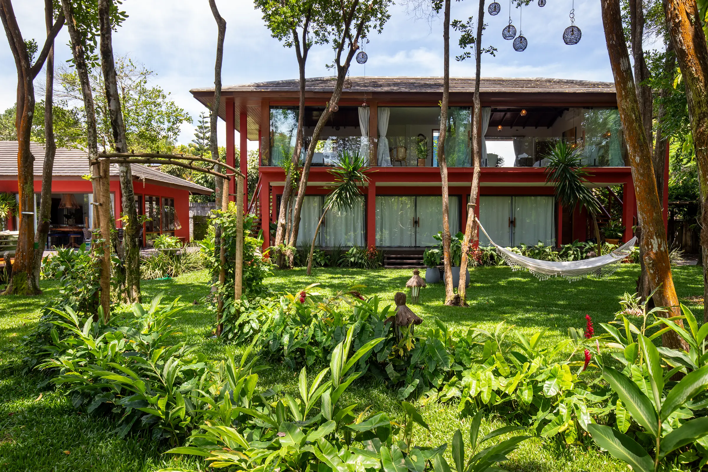 Fachada do casarão de dois andares com pilares vermelhos, vista do jardim com uma rede em primeiro plano