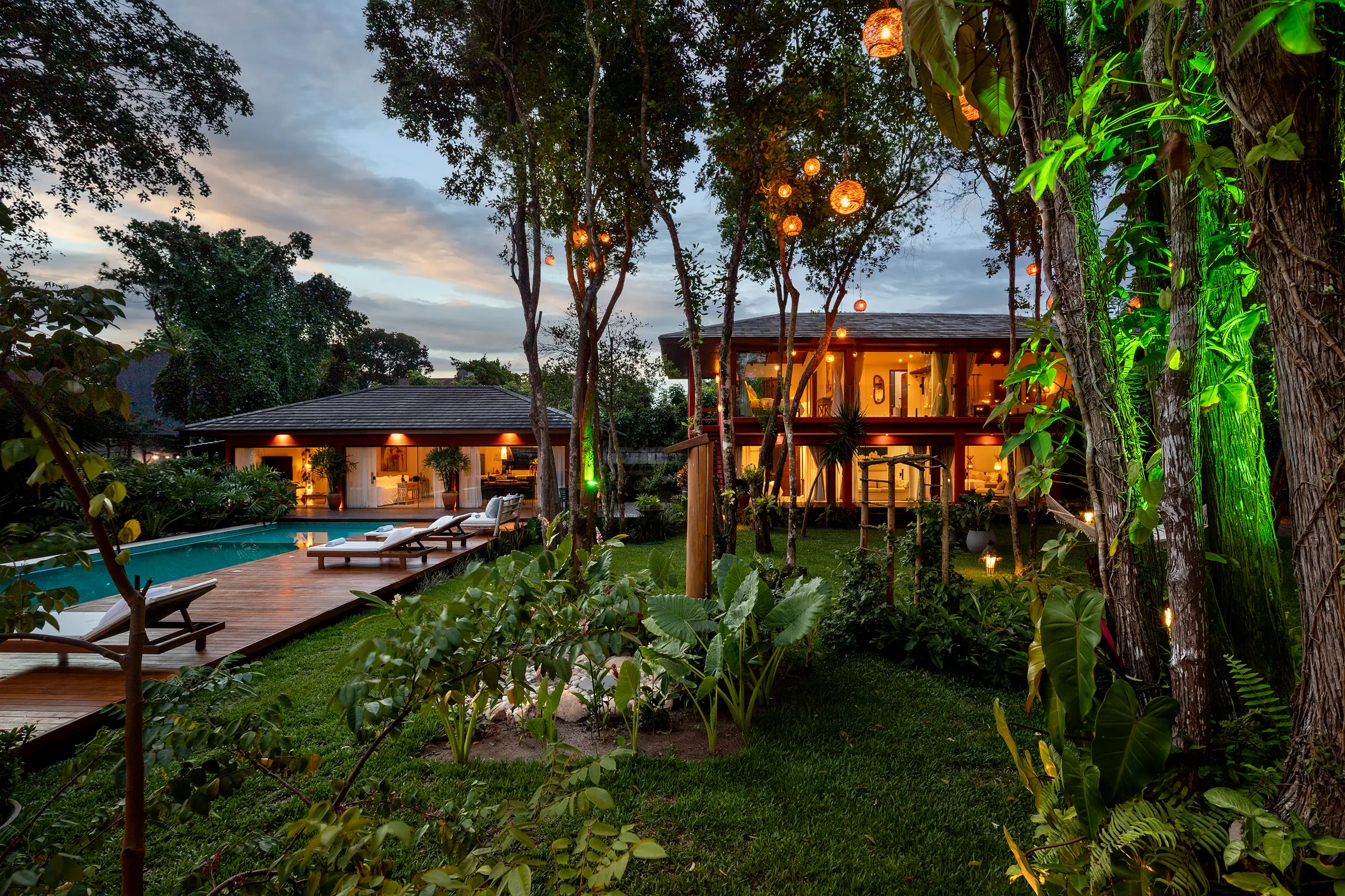 Jardim ao entardecer com a piscina, a casa da piscina e o casarão vermelho iluminados por luz verde e âmbar