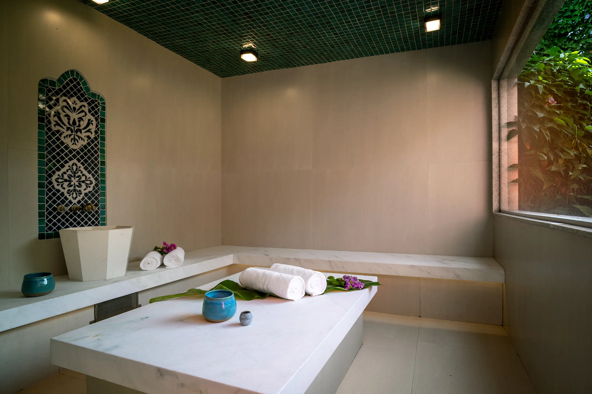 Sauna turca com bancada de mármore, nicho com fonte de mosaico, toalhas e teto de mosaico verde