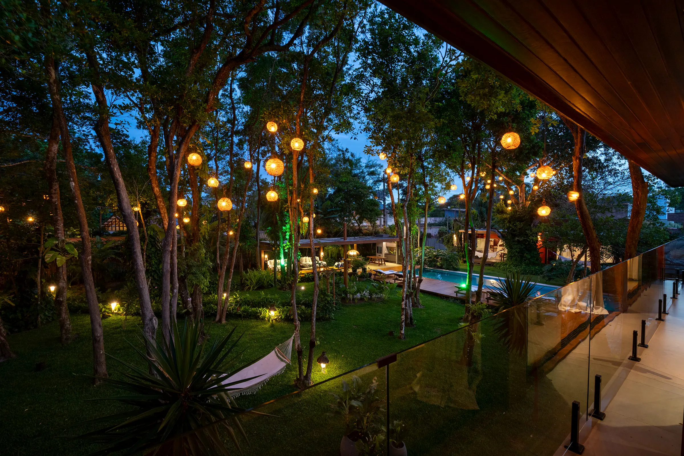 Varanda de vidro sobre o jardim iluminado, com a rede, o pavilhão e a piscina ao longe