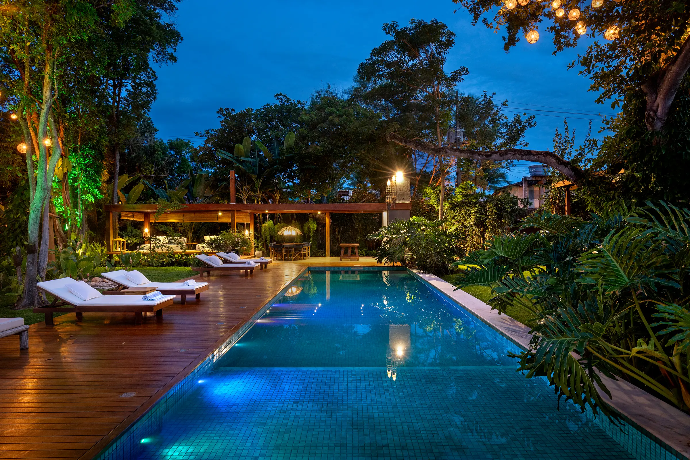 Piscina e pavilhão aberto no azul do fim de tarde, com lanternas acesas entre a vegetação