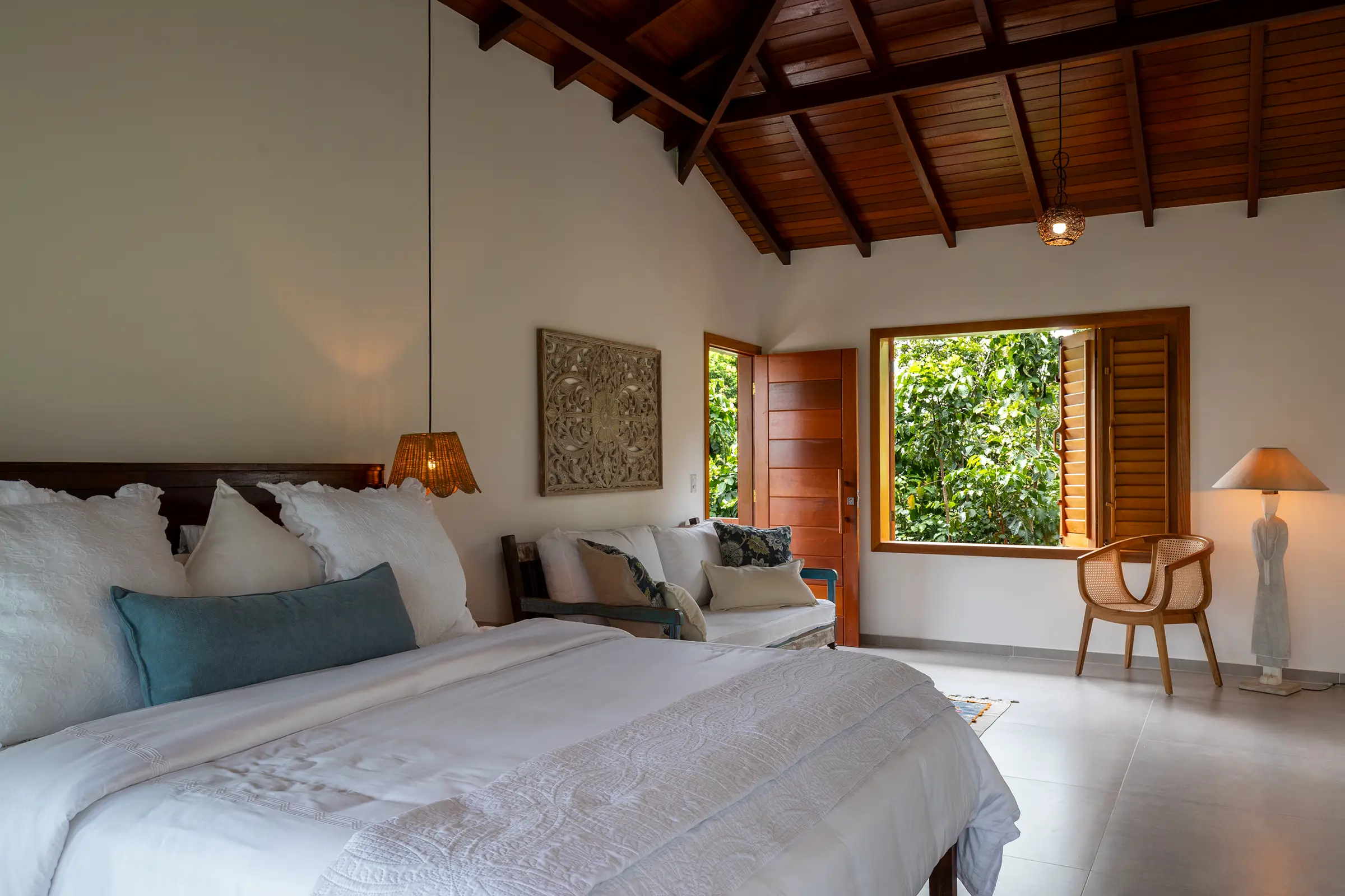 Quarto com cama de cabeceira escura, daybed, painel entalhado e janela com persianas de madeira